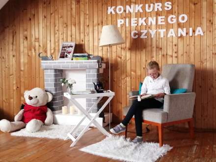 Konkurs Pięknego Czytania w SP2 Istebna; uczestniczka w fotelu z książką; w tle kominek i miś zabawka