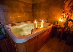 Jacuzzi; na brzegu dwie świece i dwa kieliszki, nieopodal stolik z trzema świecami, ciepłe światło, wszystko w brązach i drewnie, trzy doniczki z kwiatkami, jedna mała na stoliku, dwie duże stoją na ziemi