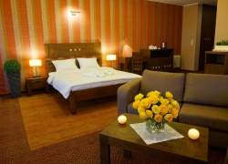 Wnętrze sypialni hotelowej, na pierwszym planie stolik z bukietem żółtych róż stojącym na białej serwetce, dale beżowa sofa, łóżko małżeńskie zasłane białą pościelą, stylowe, proste ciemnobrązowe dwie niewielkie komody; na podłodze wykładzina dywanowa - d