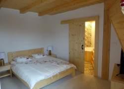 Wnętrze sypialni; białe ściany, jasne drewniane meble - łóżko małżeńskie i szafki nocne, pościel biała, na niej kwiatowy wzór, drewniane drzwi leeko odsunięte, za nimi widoczny fragment łazienki