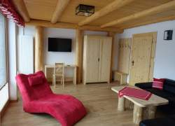 Wnętrze pokoju – strefa wypoczynku – czarna kanapa, czerwony leżak, jasne drewniane meble – stolik, szafa, takiż też drewniany sufit i drzwi; na stoliku czerwona serwetka w biały wzór