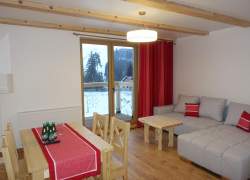 Szara kanapa narożnikowa, obszerna na niej czerwone poduszki i obok z jasnego drewna stolik, duże okno balkonowe z czerwoną kotarą, stół z jasnego drzewa i cztery krzesła, czerwony obrus, na nim szklanki i woda w małych, szklanych zielonych butelkach oraz