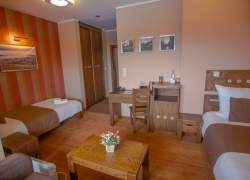 Wnętrze sypialni – dwa łóżka, szafa z przesuwanymi drzwiami, na ścianach zdjęcia. Dwie szafki nocne, dwa stoliki, krzesło. Bazowa wykładzina na podłodze. Ściana z tkaniną w rude pasy.