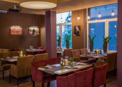 Wnętrze restauracji Bukova, widocznych pięć stolików z beżowymi wyściełanymi fotelami oraz fotelami w kolorze jasnego wina, wnętrze w beżach i brązach na ścianach stare zdjęcia; pośrodku pomieszczenia filar