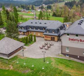 Widok obiektu z lotu ptaka – dwa proste w architekturze obiekty w tonacji szaro-beżowej z grafitowymi pokryciami dachów oraz wiata grillowa, wyłożony kostką plac, na nim sześć stolików i krzesła – ogródek  letni restauracji, wokół nieco zieleni, drzewa, z