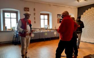 Józef Michałek i fotoreporterzy na sali wystawowej ośrodka kultury, w tle okna , stoły z eksponatami oraz po prawej Największa Koronka Koniakowska Świata