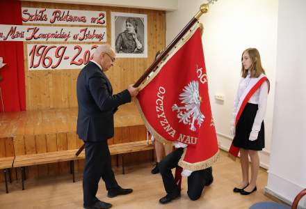 Dyrektor Szkoły Podstawowej nr 2 Bogdan Ligocki przekazuje sztandar Szkolnemu Pocztowi Sztandarowemu; sztandarowy przyklęka by go ucałować; sztandar skierowany orłem białym na czerwonym tle do publicznośc