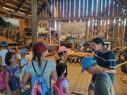 Grupa dzieci w stodole Muzeum Regionalnego Na Grapie słucha kustoszki,  wkoło dawne sprzęty rolnicze i obrazy przedstawiające krajobraz wsi sprzed lat.