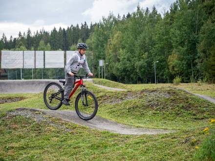 Chłopiec w kasku i szarej bluzie z numerem startowym jedzie na czerwonym rowerze górskim po torze pumptrack. W tle widać trawiaste wzniesienia, białe ekrany oraz gęsty, zielony las.