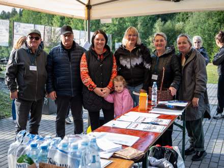 Grupa organizatorów z małą dziewczynką w różowej bluzie stoi przy stole pod namiotem. Na blacie leżą dyplomy, koszyk i dokumenty, a na pierwszym planie zgrzewki wody butelkowanej.