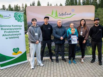 Chłopiec dumny trzyma dyplom na tle ścianki promocyjnej Istebnej, otoczony przez dorosłych i organizatorów. Po lewej stronie stoi zielono-biały baner, a w tle widać zarys lasu.