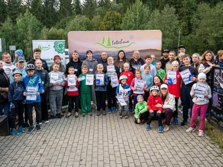 Grupowe zdjęcie pamiątkowe dzieci i dorosłych na świeżym powietrzu. Dzieci trzymają dyplomy i spoglądają w stronę obiektywu. W tle widnieje zielona ścianka reklamowa z napisem Istebna oraz malowniczy, leśny krajobraz.
