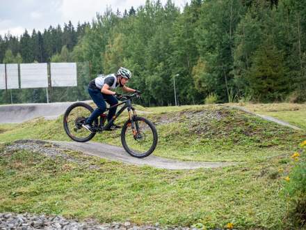 Młody kolarz w kasku i sportowym stroju z numerem startowym jedzie pochylony na czarnym rowerze górskim po torze pumptrack. W tle widać trawiaste pagórki, ogrodzenie oraz gęsty las.