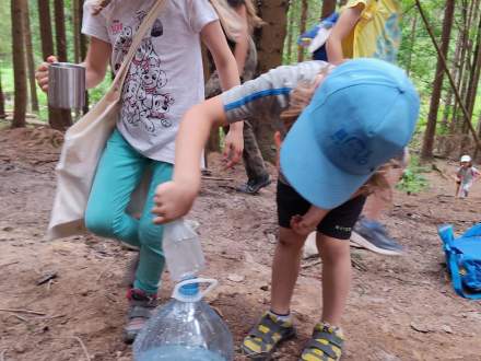 Dzieci bawiące się w lesie; dziewczynka z metalowym kubeczkiem w dłoni, chłopiec nalewa wodę do dużej butelki