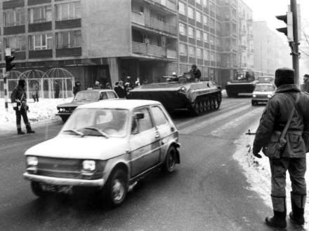 Czarno-białe zdjęcie ulicy, na której poruszają się samochody i czołgi oraz służby mundurowe; f. dzieje.pl