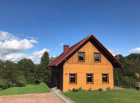 Drewniany domek widziany od szczytu – na parterze trzy okna, na piętrze dwa. Dwuspadowy dach. Po prawej niewielki taras, wkoło trawa.
