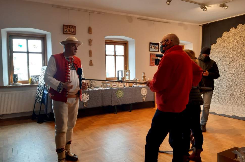 Józef Michałek i fotoreporterzy na sali wystawowej ośrodka kultury, w tle okna , stoły z eksponatami oraz po prawej Największa Koronka Koniakowska Świata