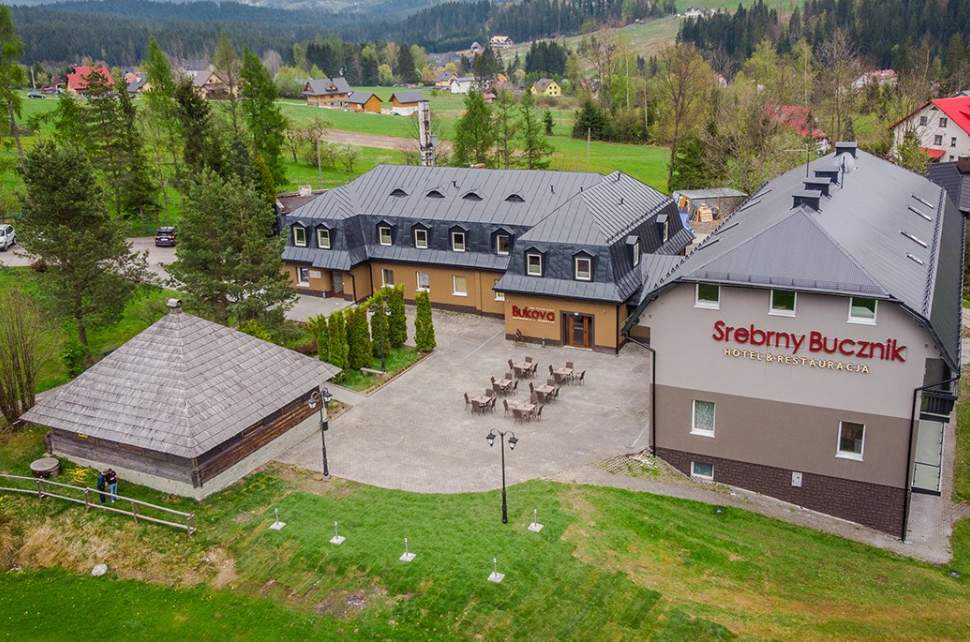 Widok obiektu z lotu ptaka – dwa proste w architekturze obiekty w tonacji szaro-beżowej z grafitowymi pokryciami dachów oraz wiata grillowa, wyłożony kostką plac, na nim sześć stolików i krzesła – ogródek letni restauracji, wokół nieco zieleni, drzewa, z