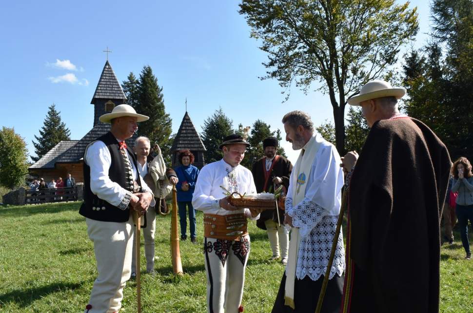 Ksiądz Proboszcz Grzegorz Kotarba odbiera bundz z rąk bacy Jana Bzdyk; Józef Michałek w guni, pasterz w czarnym brucliku, w tle kościół na Stecówce i ludzie