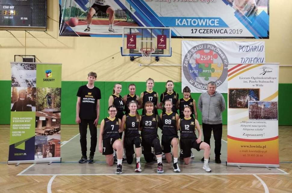 Grupa 10 uczennic w czarnych strojach sportowych z dwoma opiekunami na sali gimnastycznej; ustawione do zdjęcia w dwóch rzędach, wokół rozstawione oraz wiszące banery promocyjne, rollup szkoły i wyciągu Skolnity