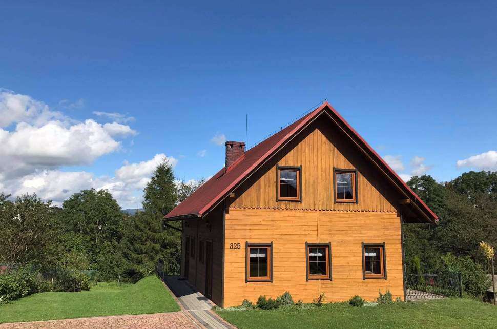 Drewniany domek widziany od szczytu – na parterze trzy okna, na piętrze dwa. Dwuspadowy dach. Po prawej niewielki taras, wkoło trawa.